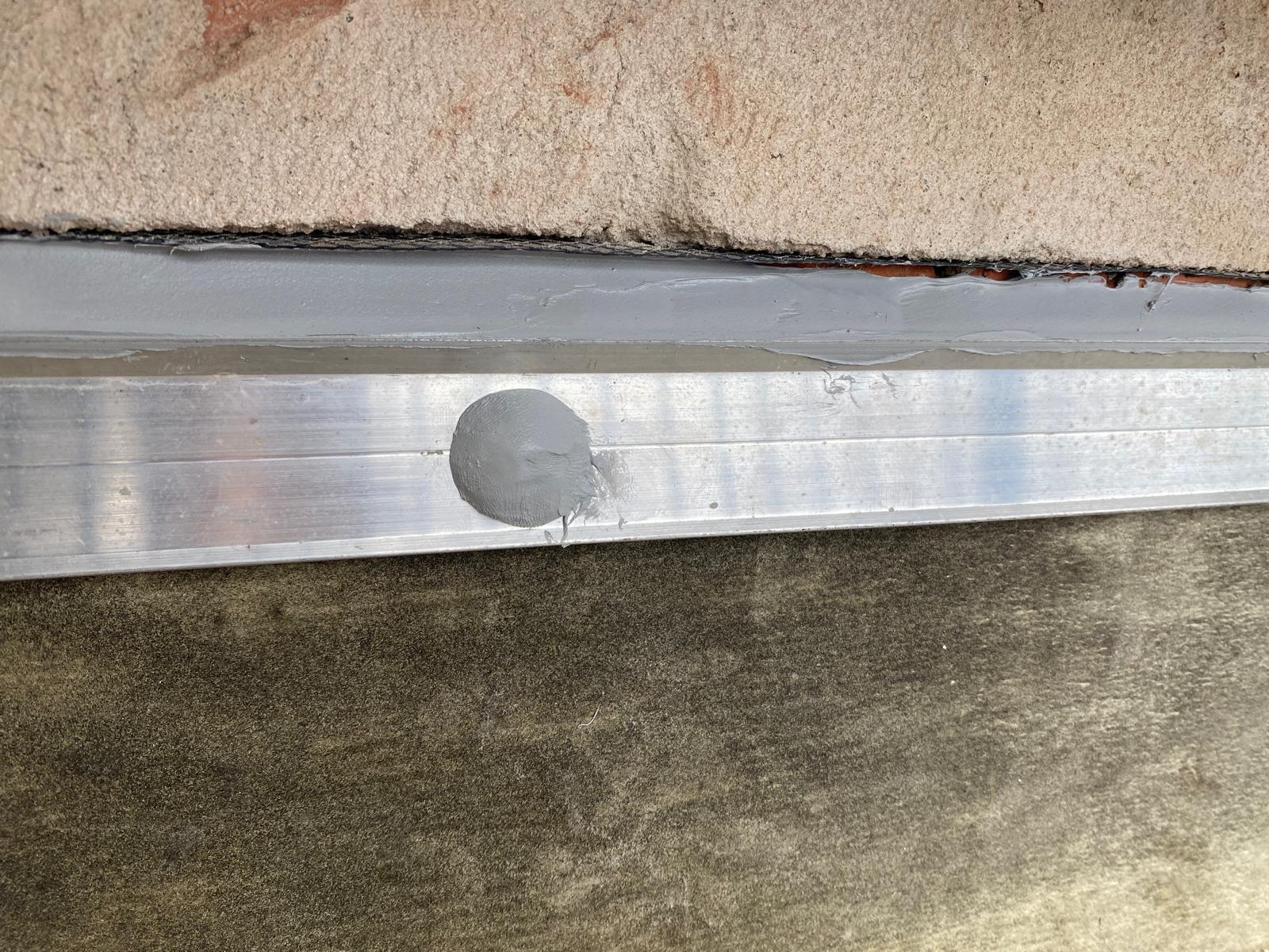 Close-up of aluminium cavity flashing strip dressed against brickwork with grey sealant bead and bolt visible